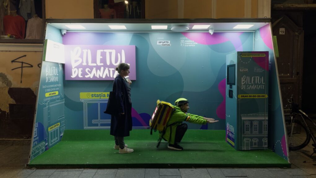 Kontinental ’25 (2025) | Crítica de la nueva película de Radu Jude | CineFilm Escena de Kontinental ’25 (2025), de Radu Jude, con una mujer esperando mientras un repartidor se agacha ante una máquina burocrática en la calle