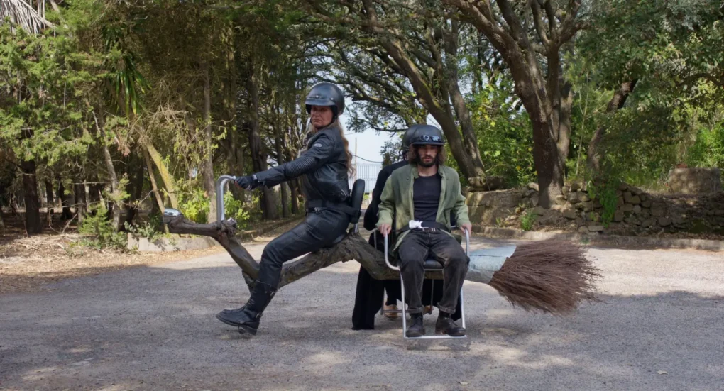 Escena de Arvore do Conhecimento (2025) con dos personajes montados en una escoba mecánica en un camino rodeado de árboles.