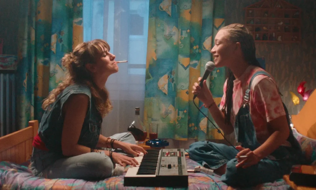 Escena de Stereo Girls (2025) con dos jóvenes protagonistas cantando y tocando un teclado en una habitación.