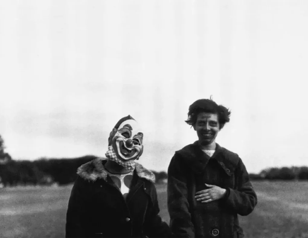 Fotografía en blanco y negro de Diane Arbus con una figura que lleva una máscara de payaso junto a otra persona al aire libre.