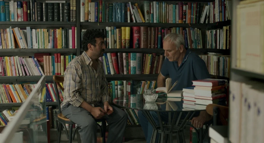 Dos hombres conversan sentados en una librería en Un simple accidente (2025), de Jafar Panahi
